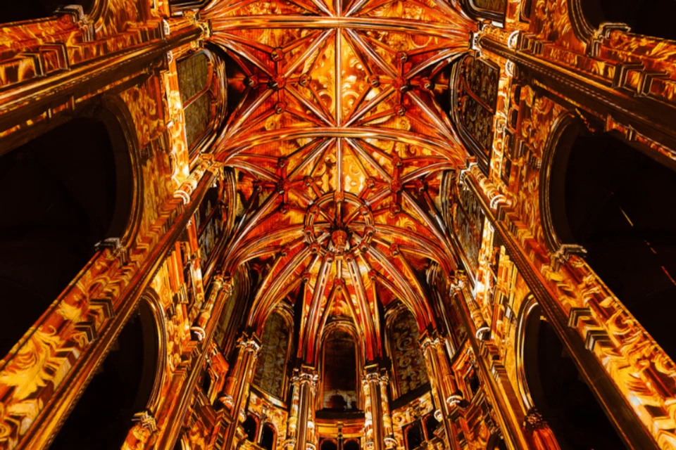 A detailed, ornate cathedral ceiling with intricate wooden beams, arches, and decorative elements illuminated warmly.