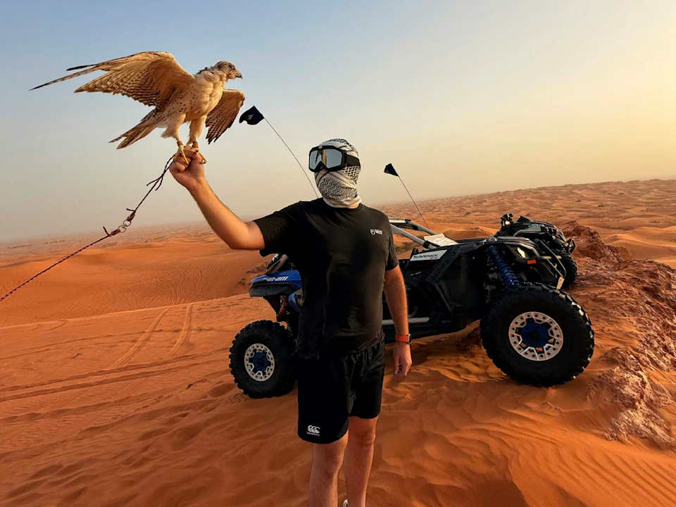 A person stands in a desert holding a falcon. An off-road vehicle with sand tires is parked nearby.
