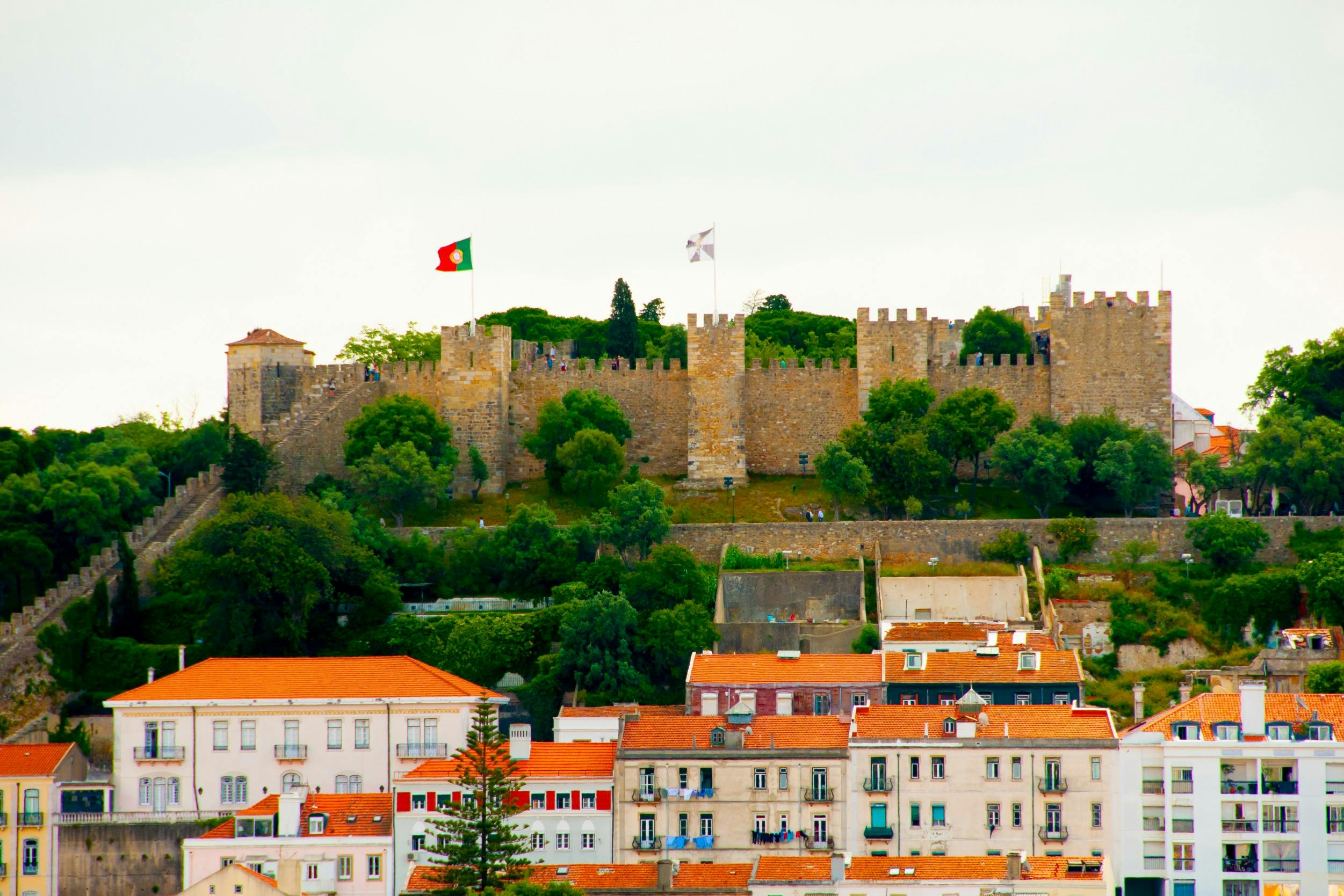 A hillside view of a historic stone castle with flags on top, surrounded by trees, above colorful houses with red roofs.
