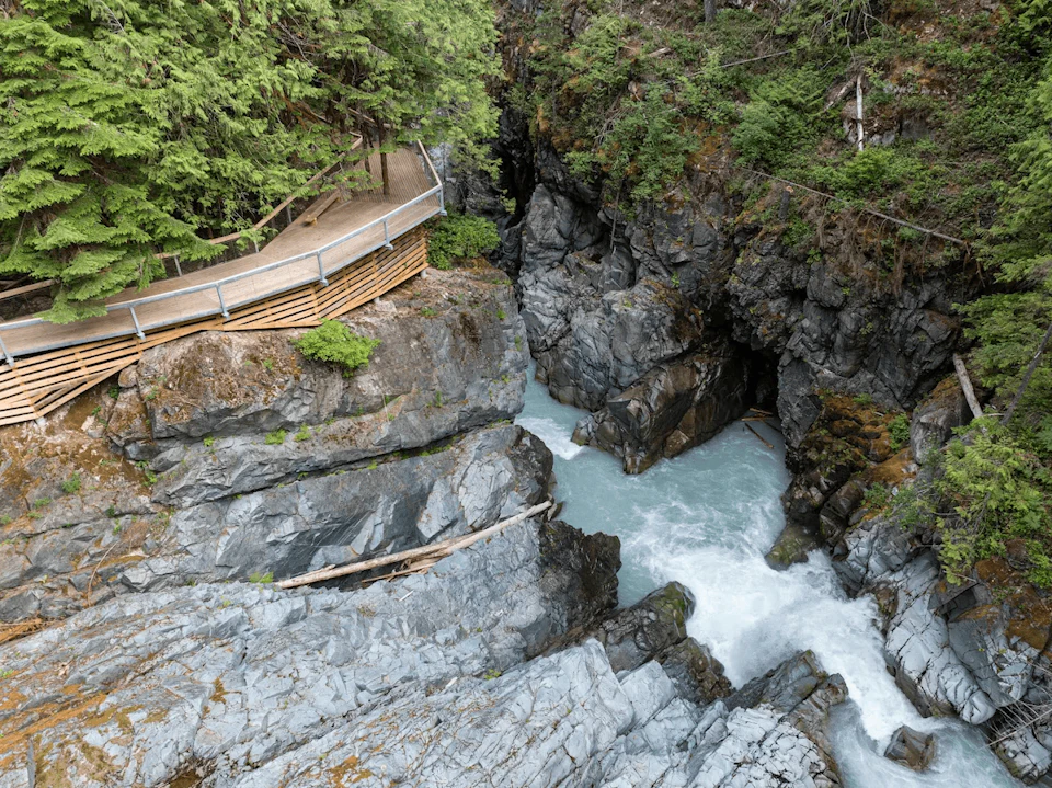 Wooden walkway with railing above a rocky gorge with a flowing river, surrounded by lush green forest.