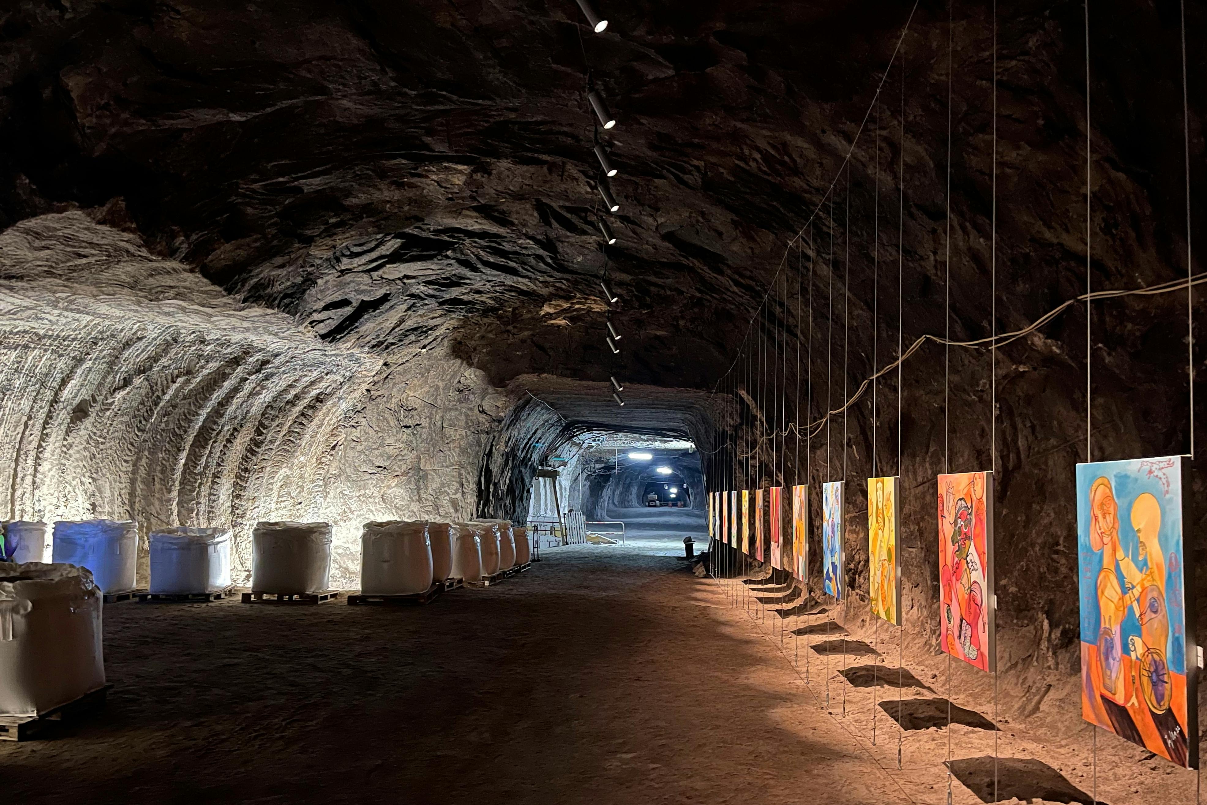A dimly lit tunnel with rock walls showcasing colorful paintings suspended on the right and large white barrels on the left.