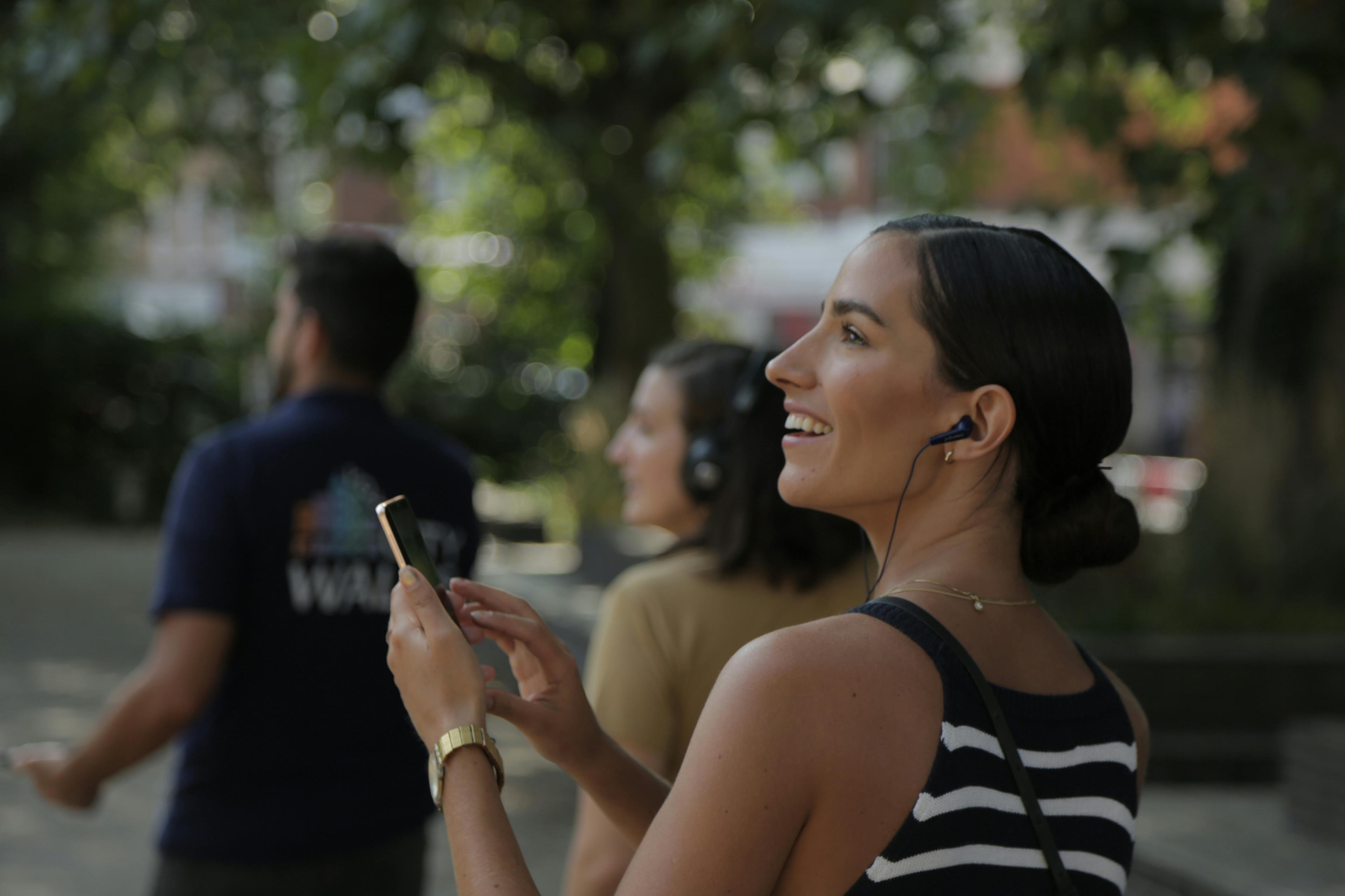 Woman with earbuds smiling, holding a smartphone in a park; two blurred people in the background also wearing headphones.