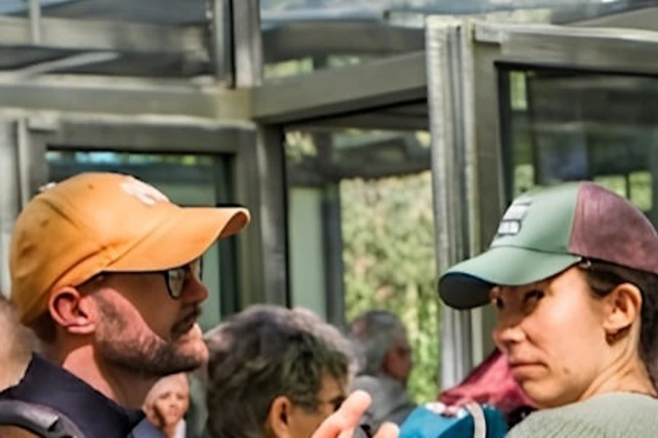 People gathered in a line, outdoors, with a man gesturing in the foreground. Glass structures and trees are visible.