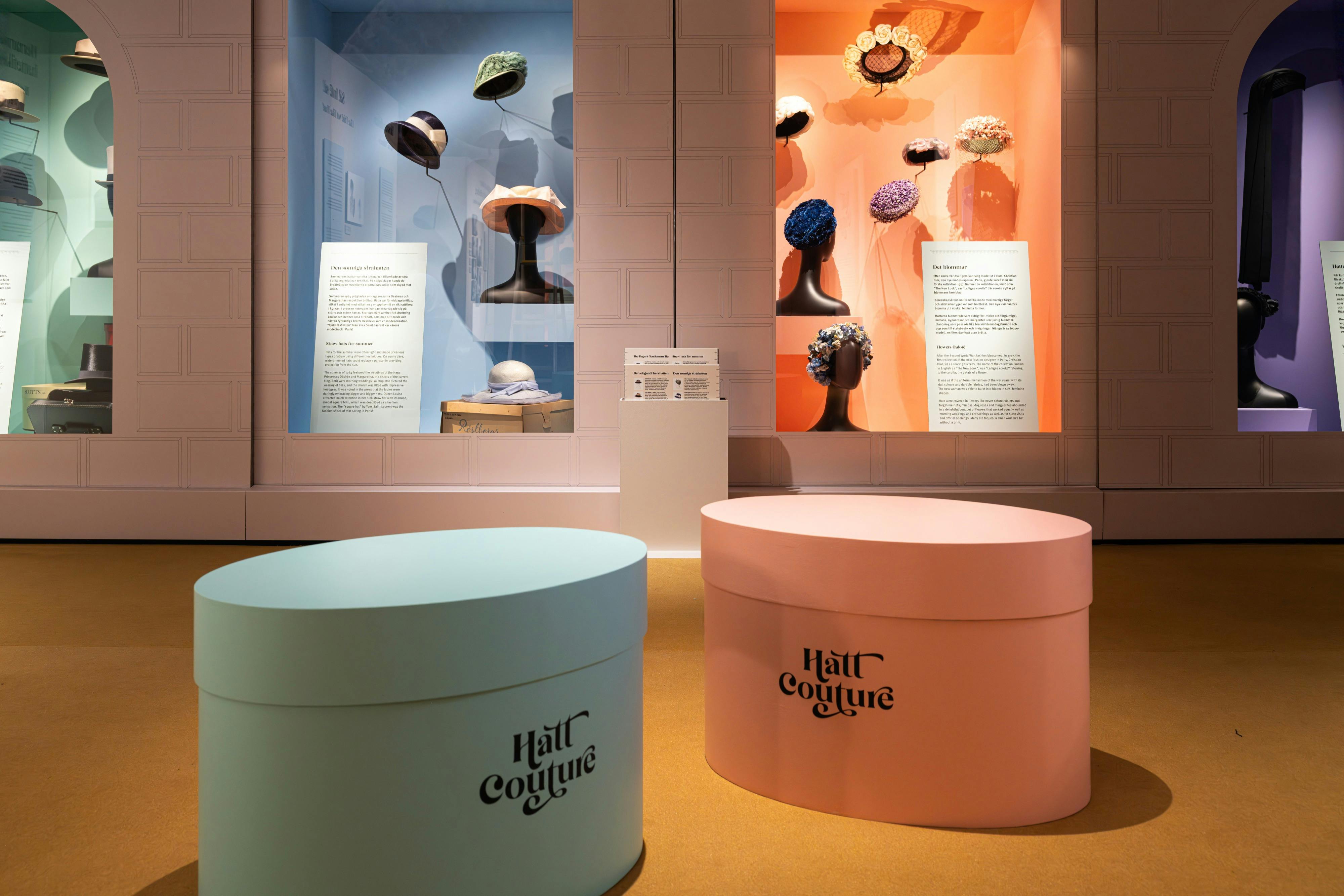 Esposizione di cappelli d'epoca in teche di vetro con testi descrittivi, abbinati a grandi cappelliere color pastello in primo piano.