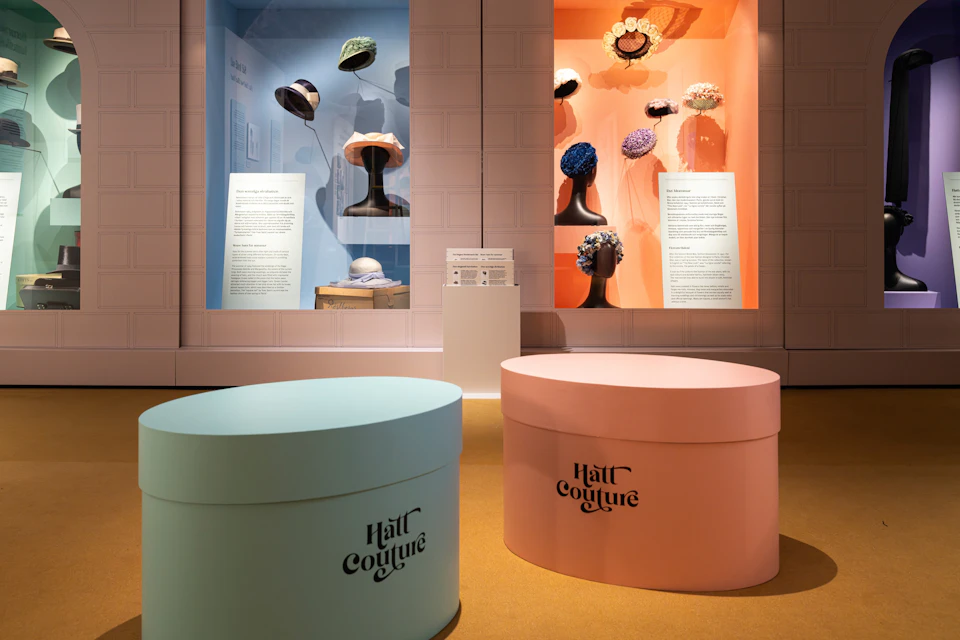 Display of vintage hats in glass cases with descriptive texts, paired with large pastel-colored hat boxes in the foreground.