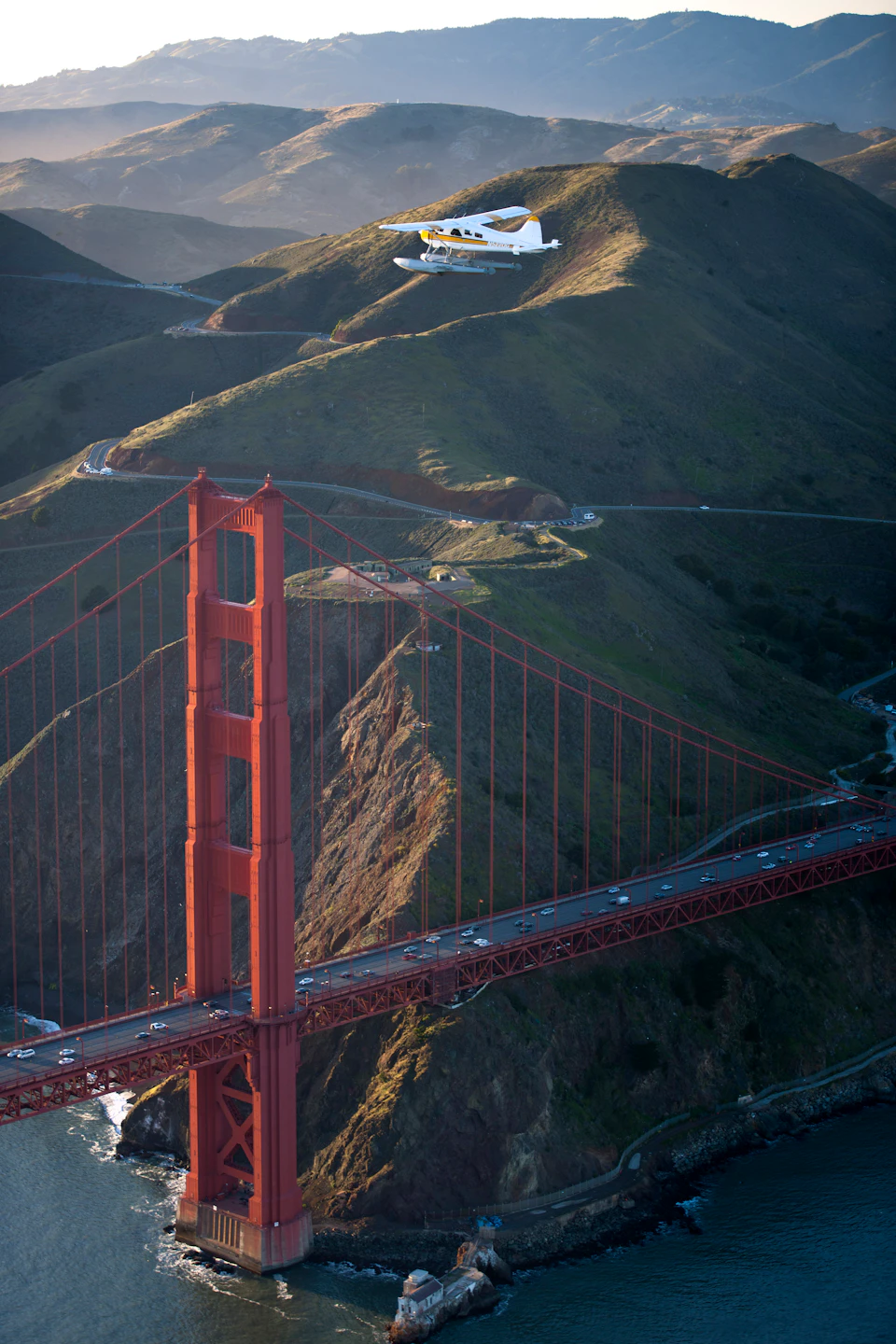 A seaplane flies over the Golden Gate Bridge with hills in the background. Cars are visible on the bridge below.