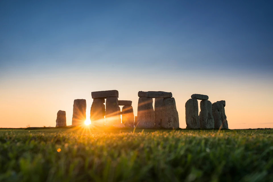 Stonehenge at sunrise with the sun casting light through the stones, set on a green field under a clear sky.