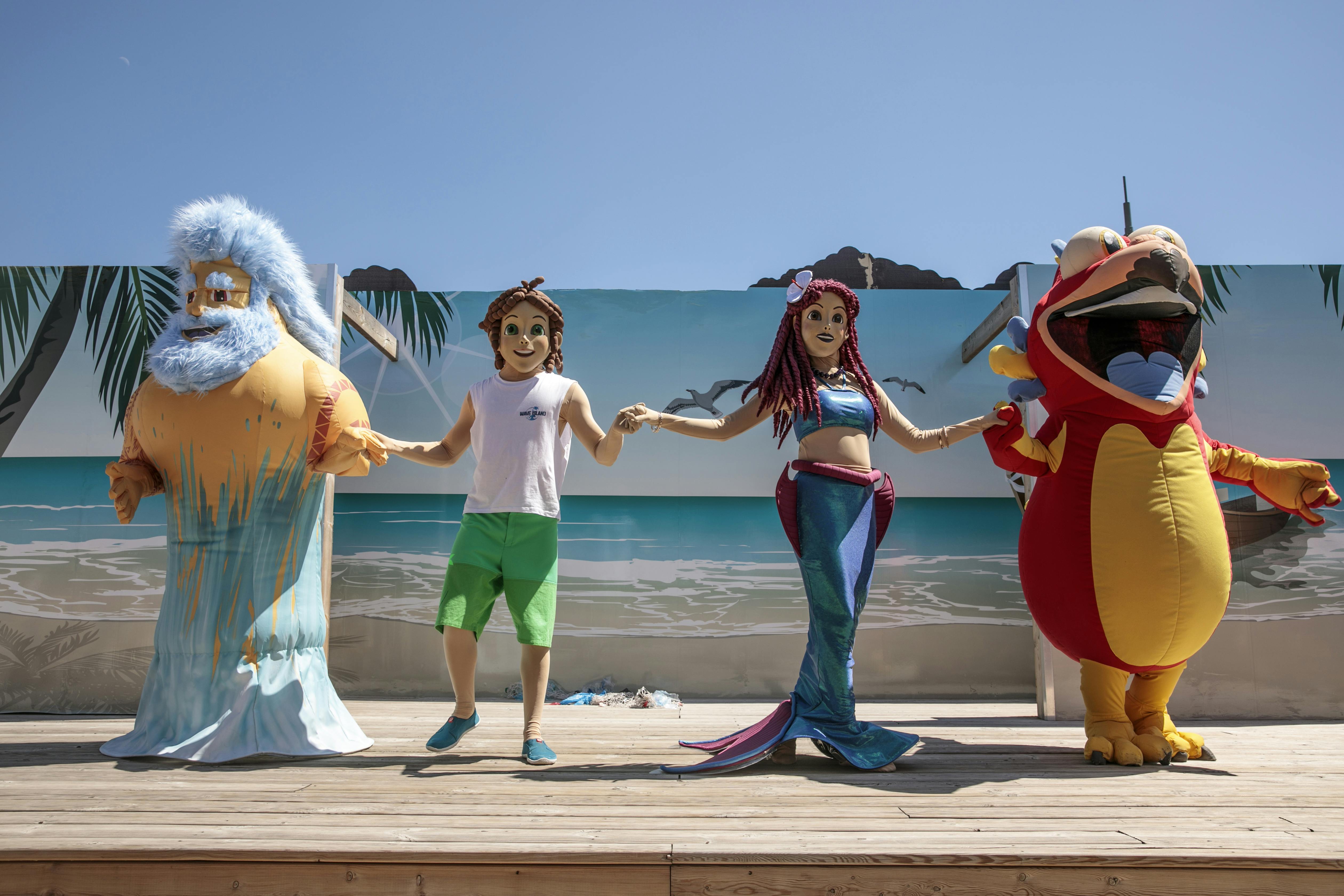 Three people in character costumes (a bearded man, a boy, and a mermaid) hold hands on a stage with a beach backdrop.