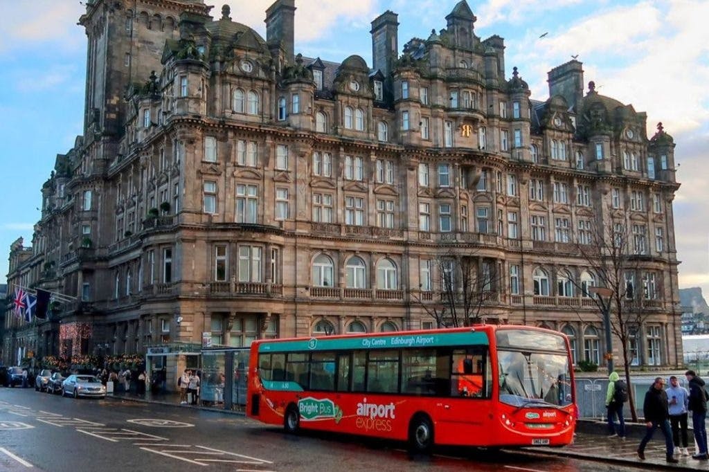 在多云的天空下，一辆红色的机场快线巴士停在一座历史悠久的华丽石头建筑附近，附近有行人和停放的汽车。