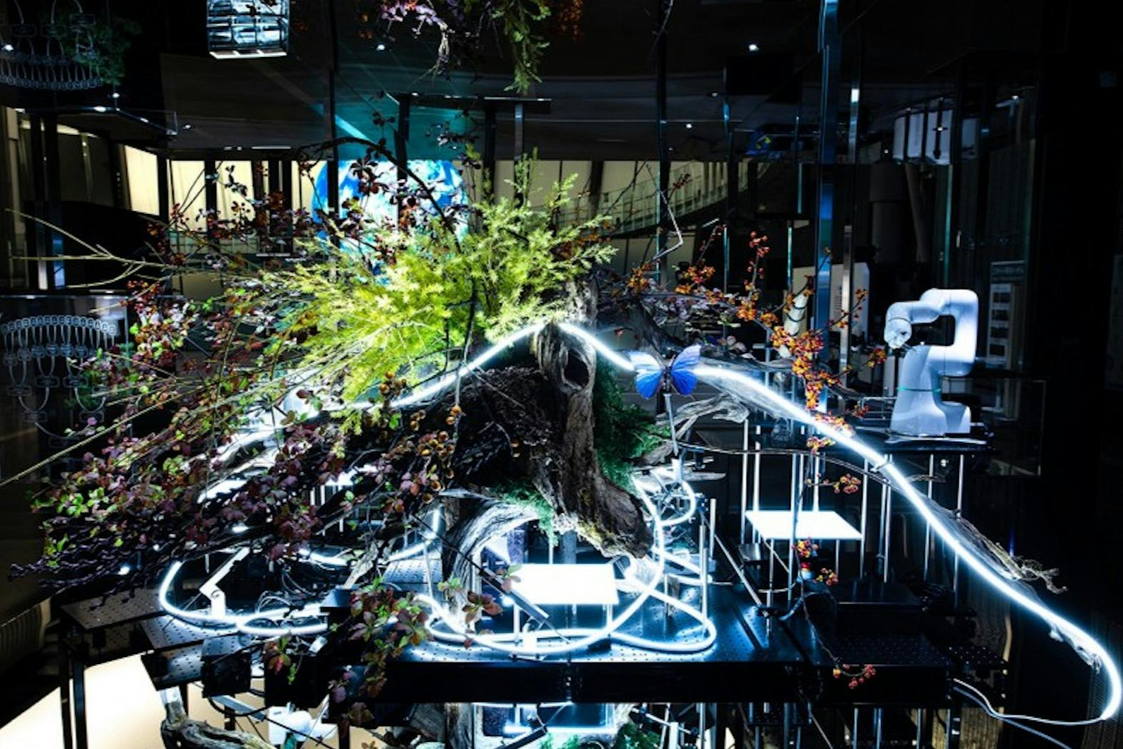 Una instal·lació d'art modern amb plantes, un braç robòtic i elements de neó brillants en un espai poc il·luminat.