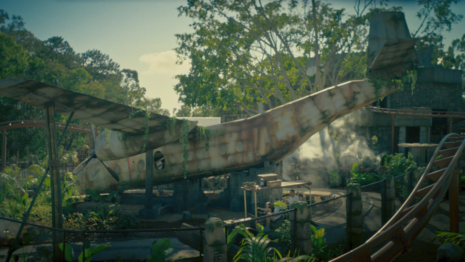 A rusted airplane fuselage surrounded by lush greenery and a small outdoor seating area, with trees and sunlight in the background.