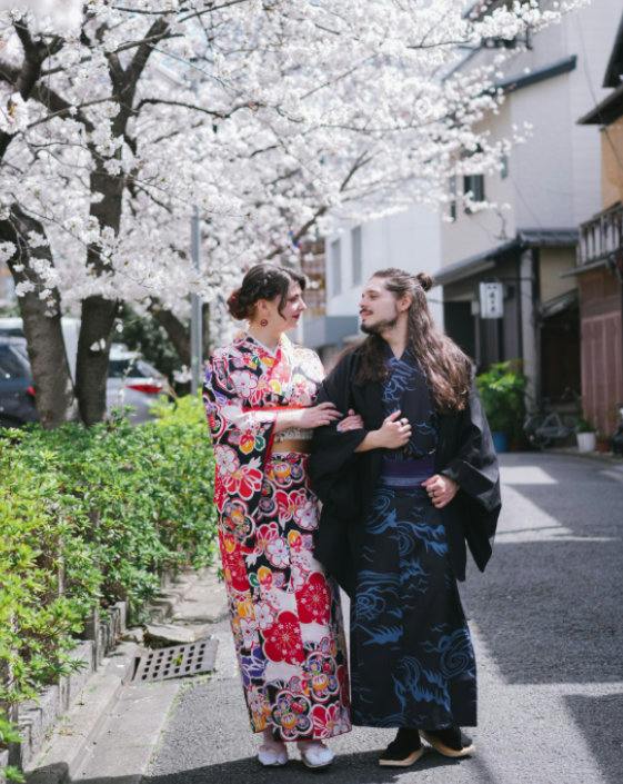 Two people in traditional Japanese attire walking beside blooming cherry trees in an urban setting.
