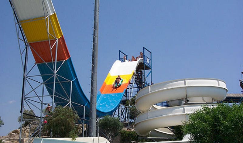 맑은 날 하얀 나선형 워터슬라이드 옆에 가파르고 다채로운 색상의 워터슬라이드를 타고 내려오는 사람들.