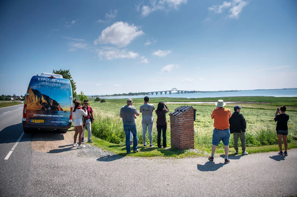 A group of people standing near a parked van, observing a scenic landscape with a bridge over water under a clear sky.