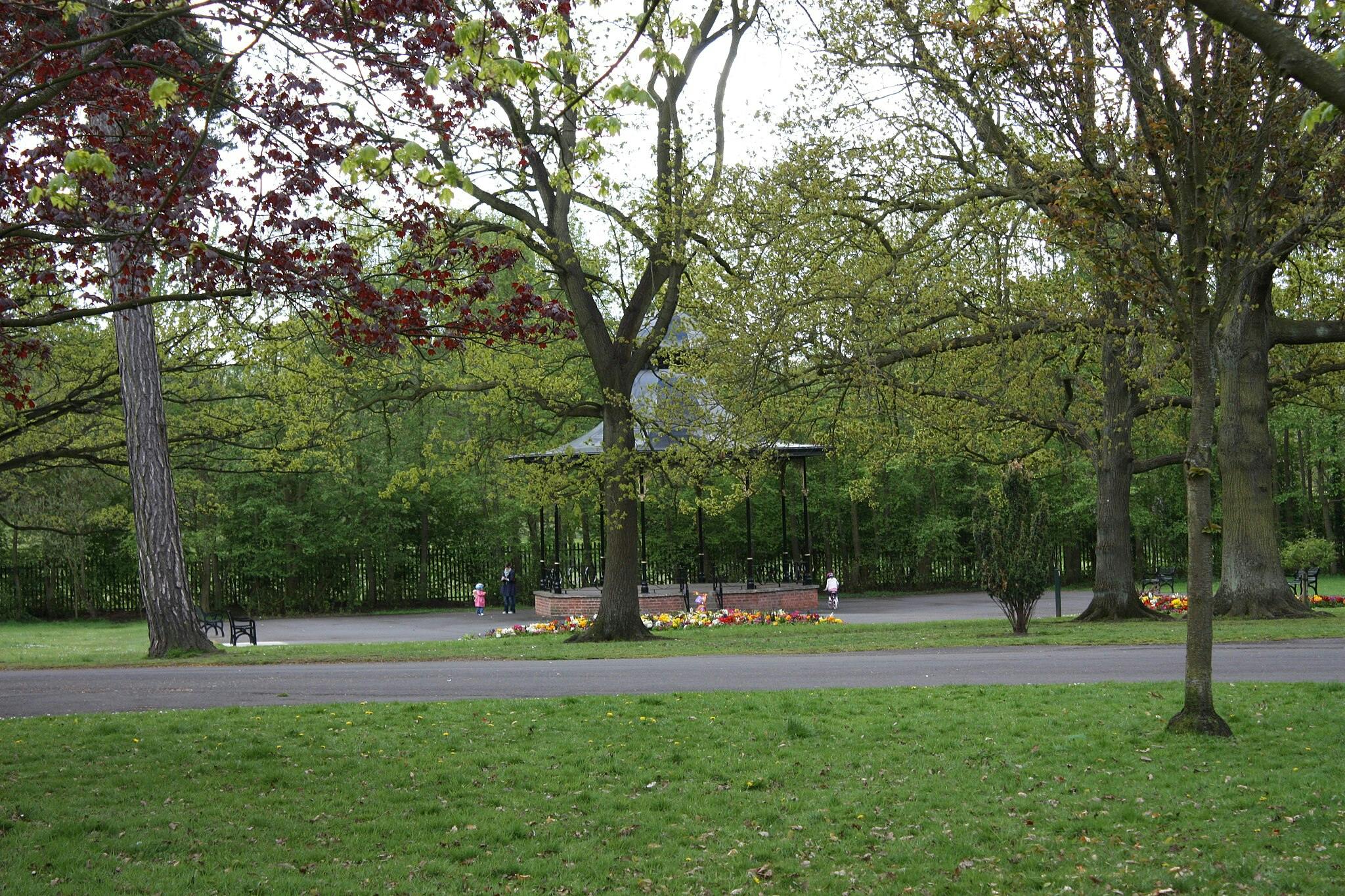 Un parco con alberi, panchine, aiuole
aiuole orali, un sentiero e un gazebo sullo sfondo. Diverse persone stanno passeggiando nelle vicinanze.