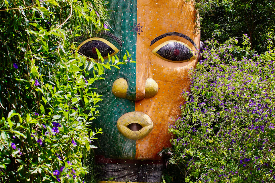 A colorful mosaic mask sculpture surrounded by lush green foliage and purple flowers.