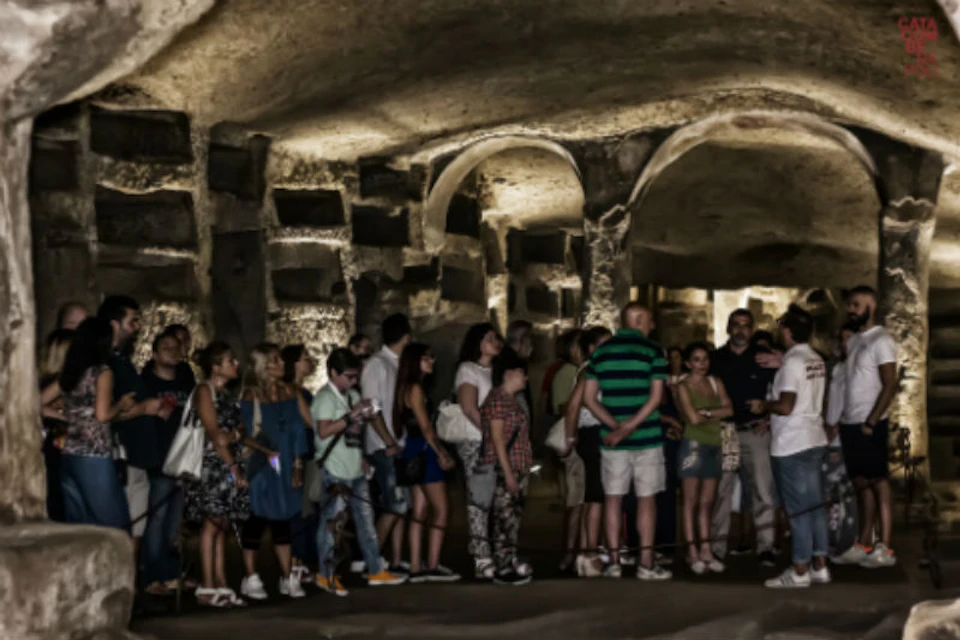A group of people stand inside an ancient, dimly lit underground structure with arches and wall niches.