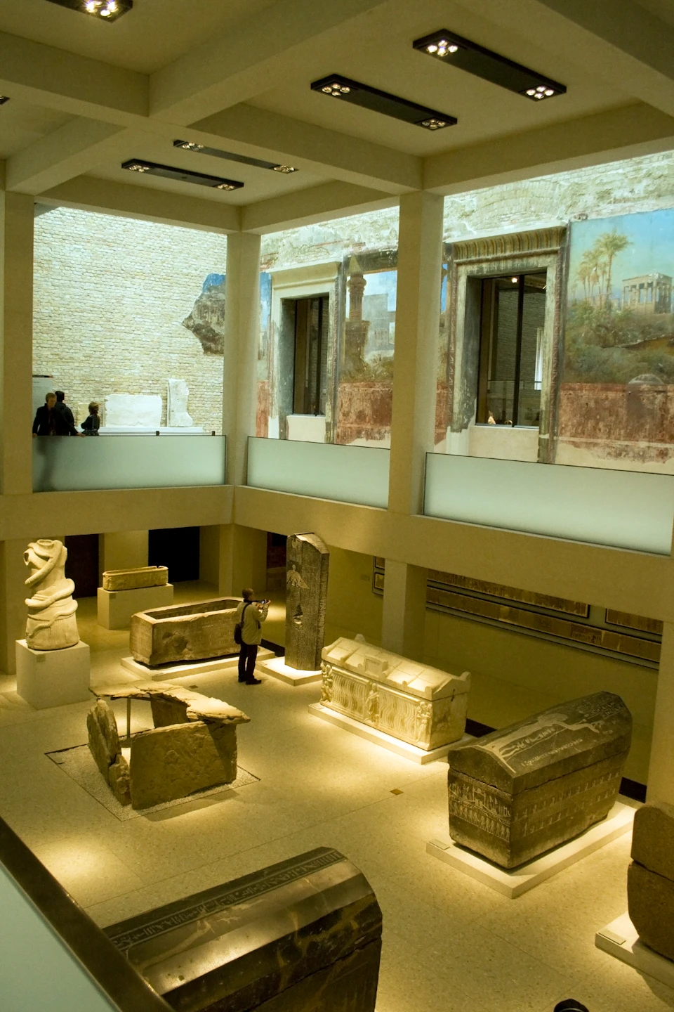 A museum exhibit with ancient sculptures, sarcophagi, and wall frescoes. Visitors observe from both ground and upper levels.