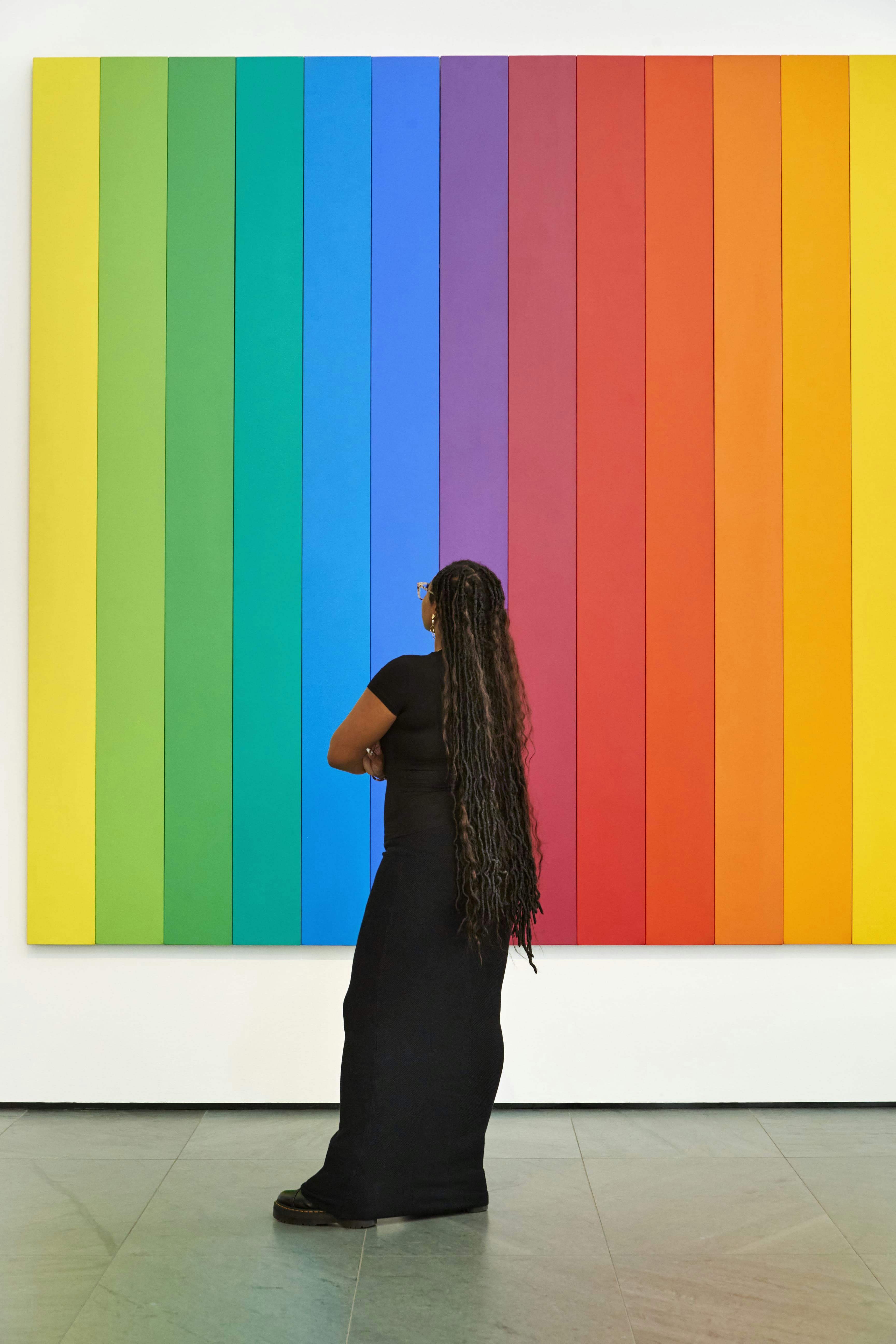 Osoba s dlouhými vlasy hledí na velké duhové pruhované umělecké dílo na bílé stěně.