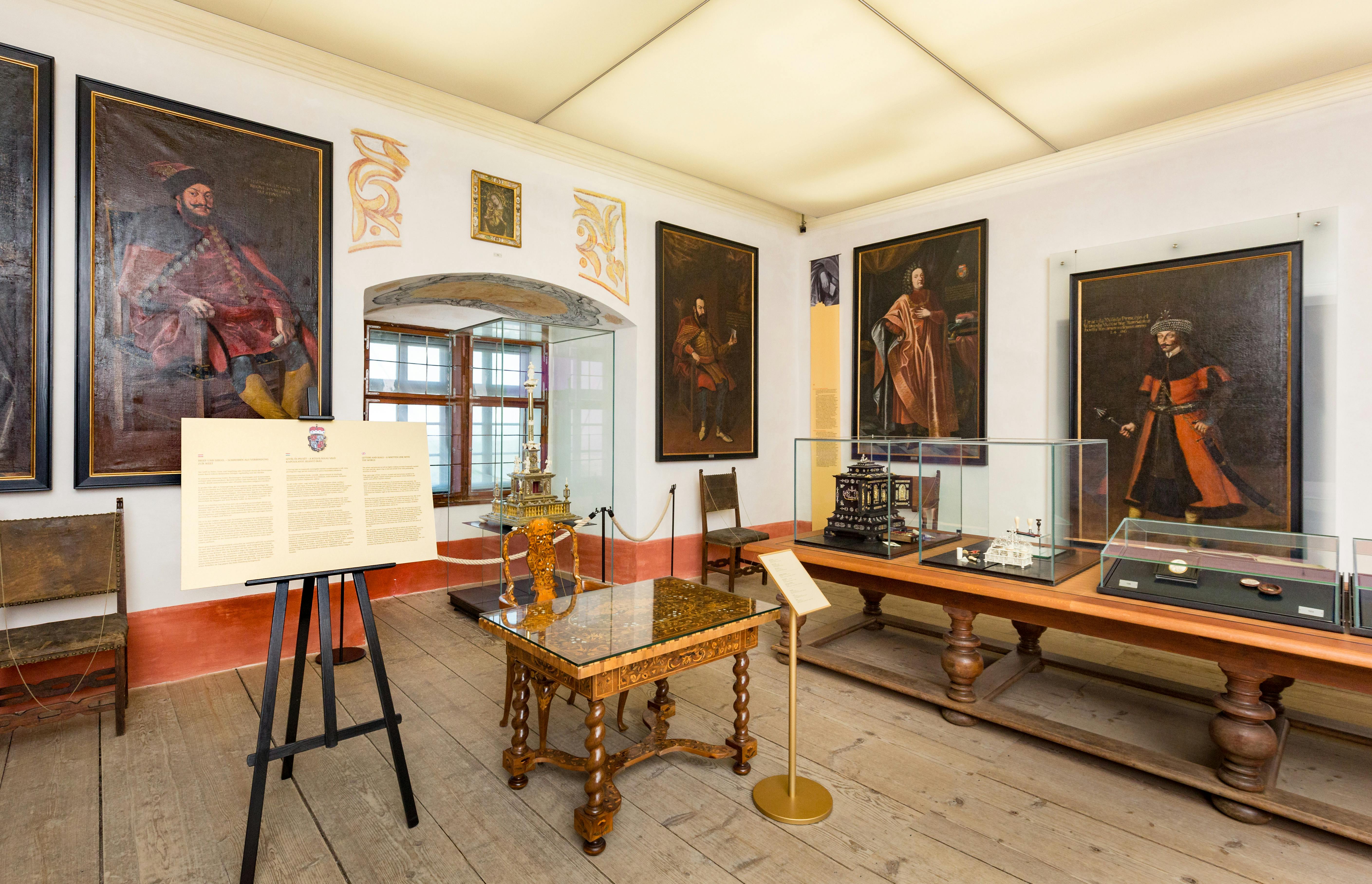 一间带有框架肖像的博物馆房间，陈列柜内装有复杂的模型、华丽的桌子和信息板。