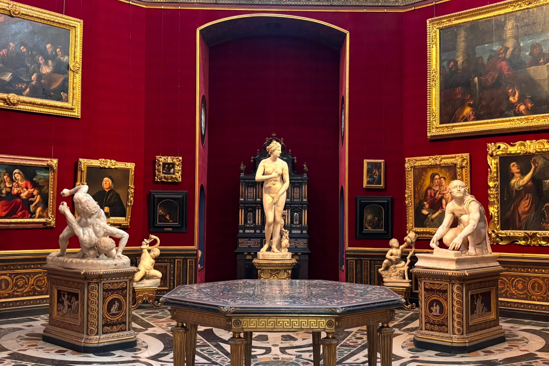 A museum gallery with red walls, ornate sculptures, framed paintings, and a decorative table in the center.