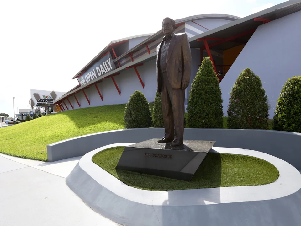Bronze statue of a man in a suit on a pedestal, in front of a building with a sign that reads "OPEN DAILY," surrounded by shrubs.