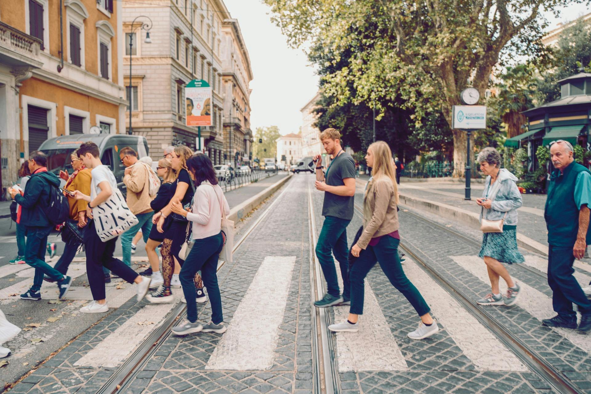 Persone che attraversano una strada alberata sulle strisce pedonali, circondate da edifici, biciclette e un orologio.