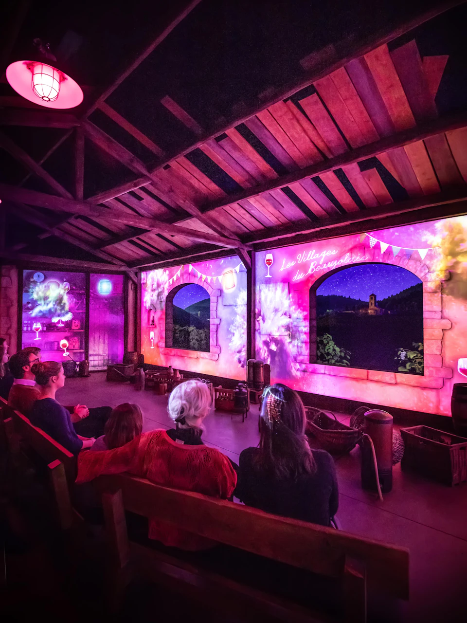 People sitting in a dimly-lit room watching colorful projected scenes on walls resembling windows, under a wooden beamed ceiling.