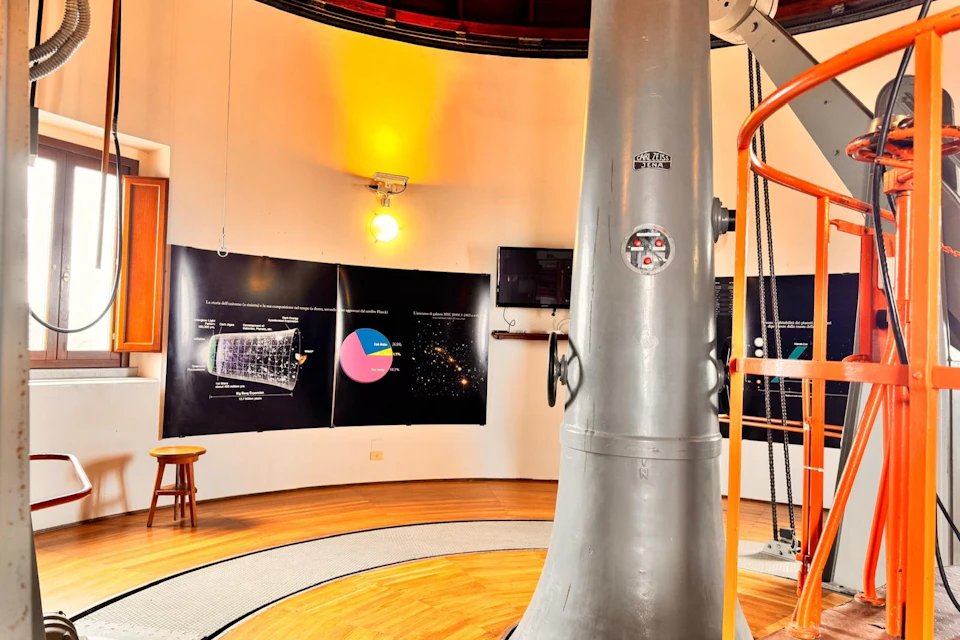 A large telescope stands in a circular room with informative posters and a yellow light on the wall.