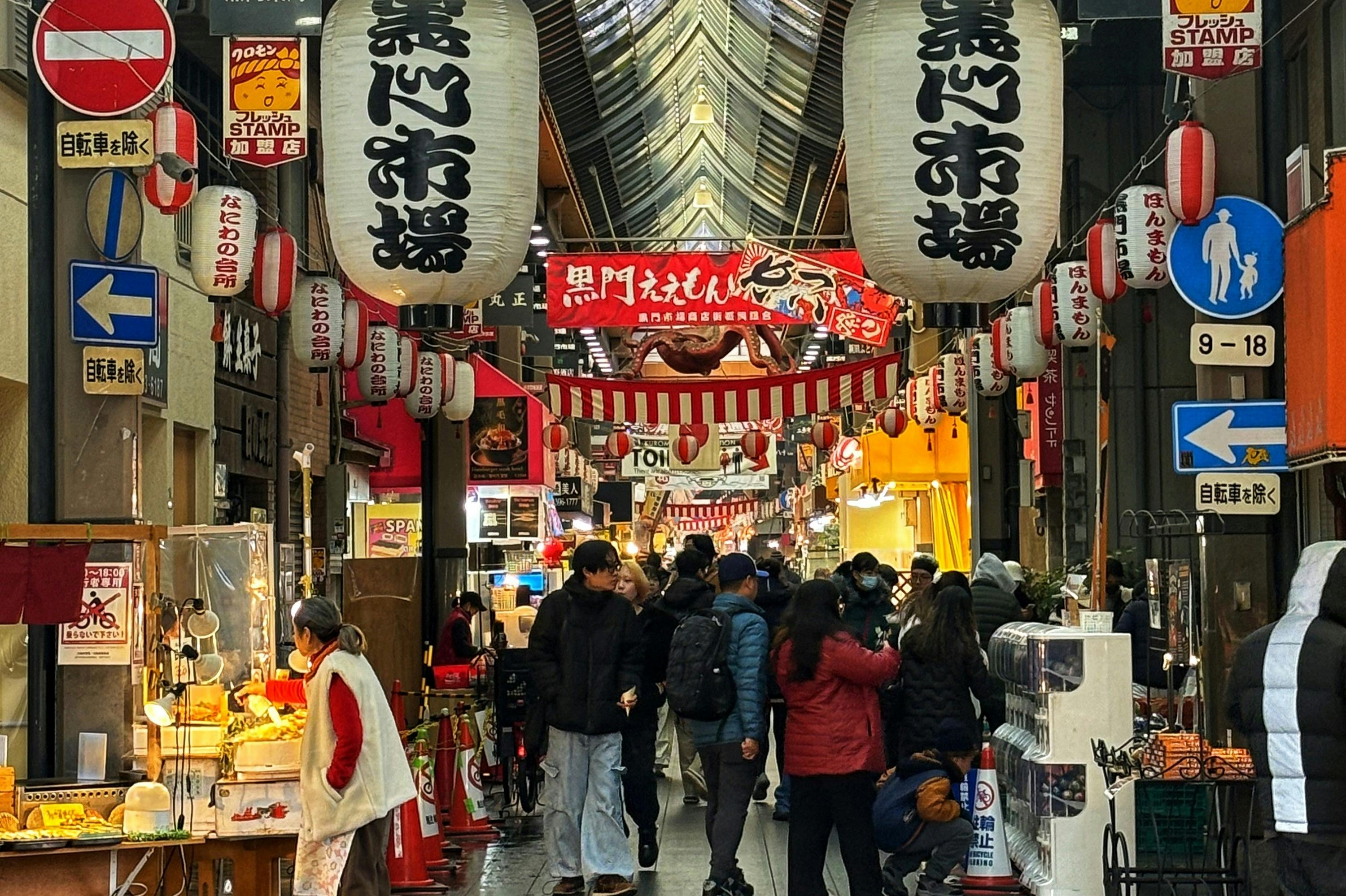 Travl japansk markedsgade med store hængende lanterner, forskellige butikker på begge sider og folk, der går og shopper.