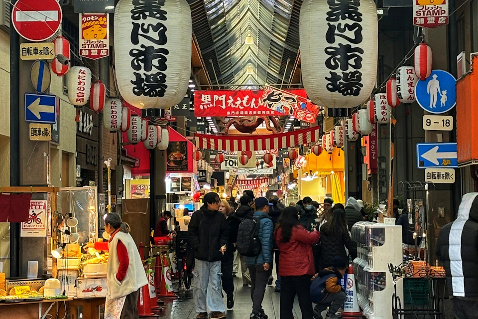 Bustling Japanese market street with large hanging lanterns, various shops on both sides, and people walking and shopping.