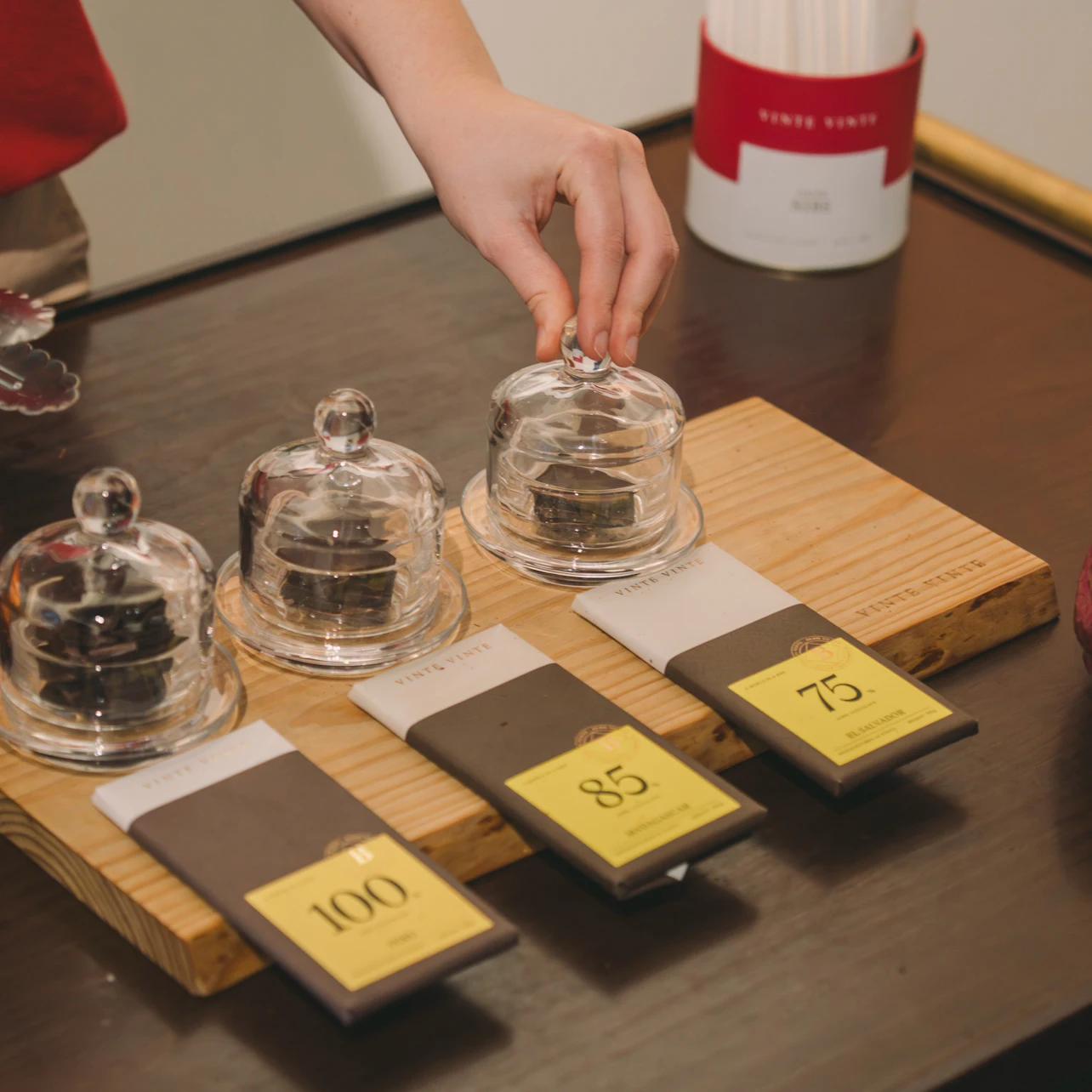 A hand reaches toward a small glass cloche covering an item, next to three chocolate bars labeled 100, 85, and 75.