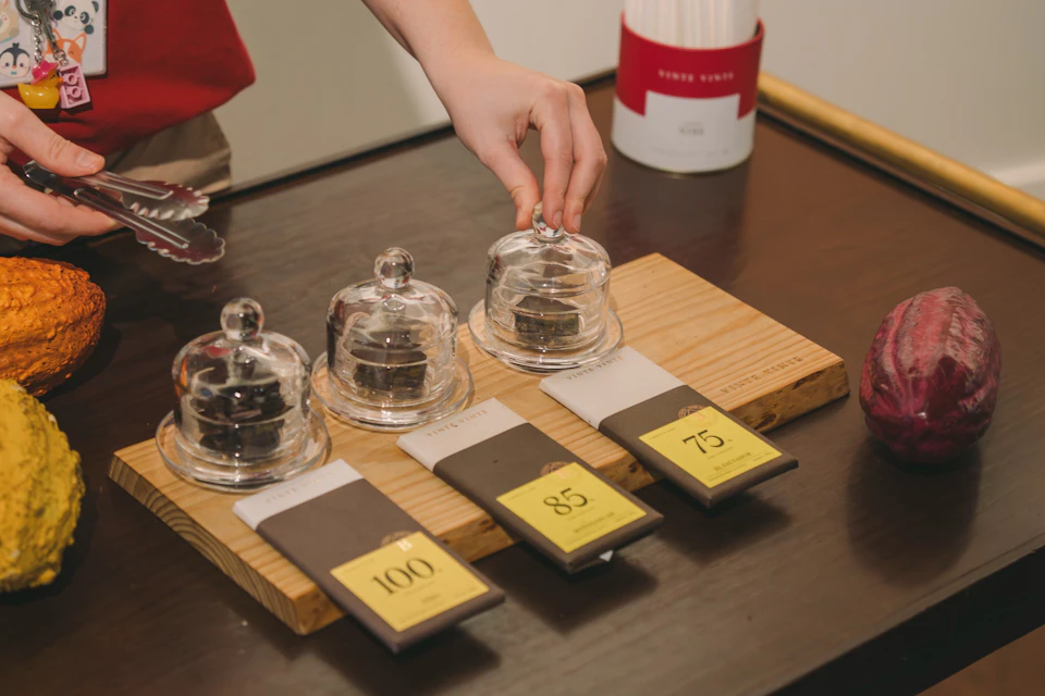 A hand reaches toward a small glass cloche covering an item, next to three chocolate bars labeled 100, 85, and 75.