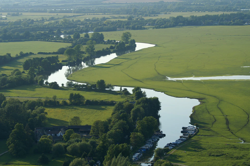 Aerial view of a winding river through green fields with trees and parked boats along the riverbank.