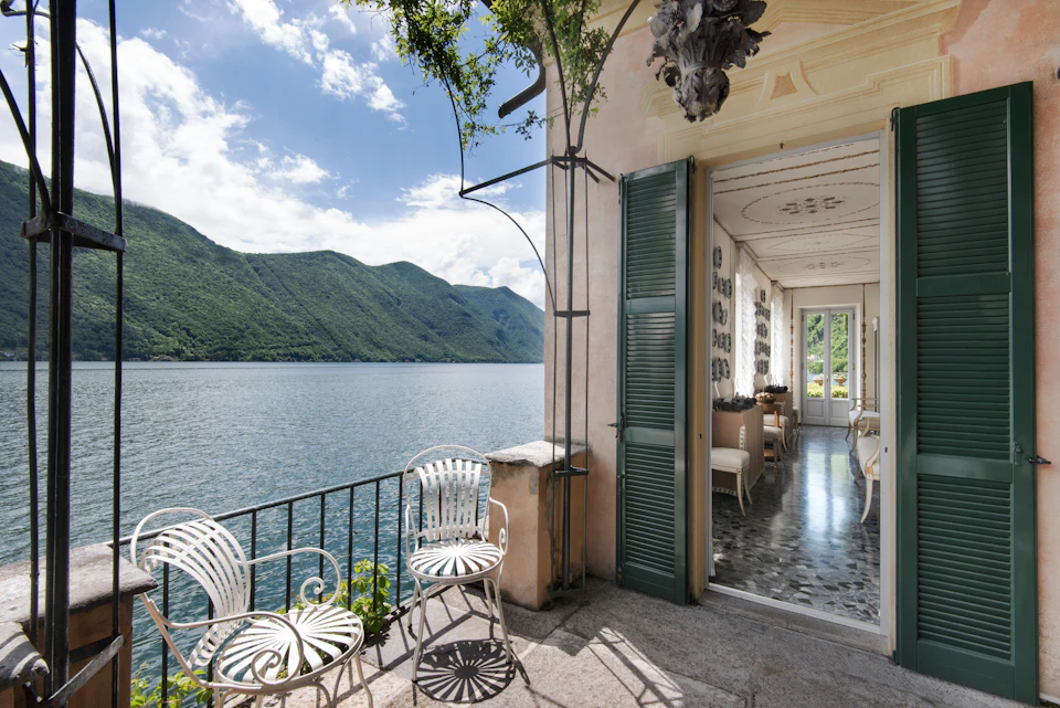 Balcony with white iron chairs overlooking a lake and green mountains, next to an open door revealing an elegant interior.