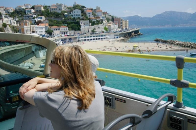 En kvinna som sitter på en turistbuss tittar på en kuststad med en strand, klart blått vatten och kullar i bakgrunden.