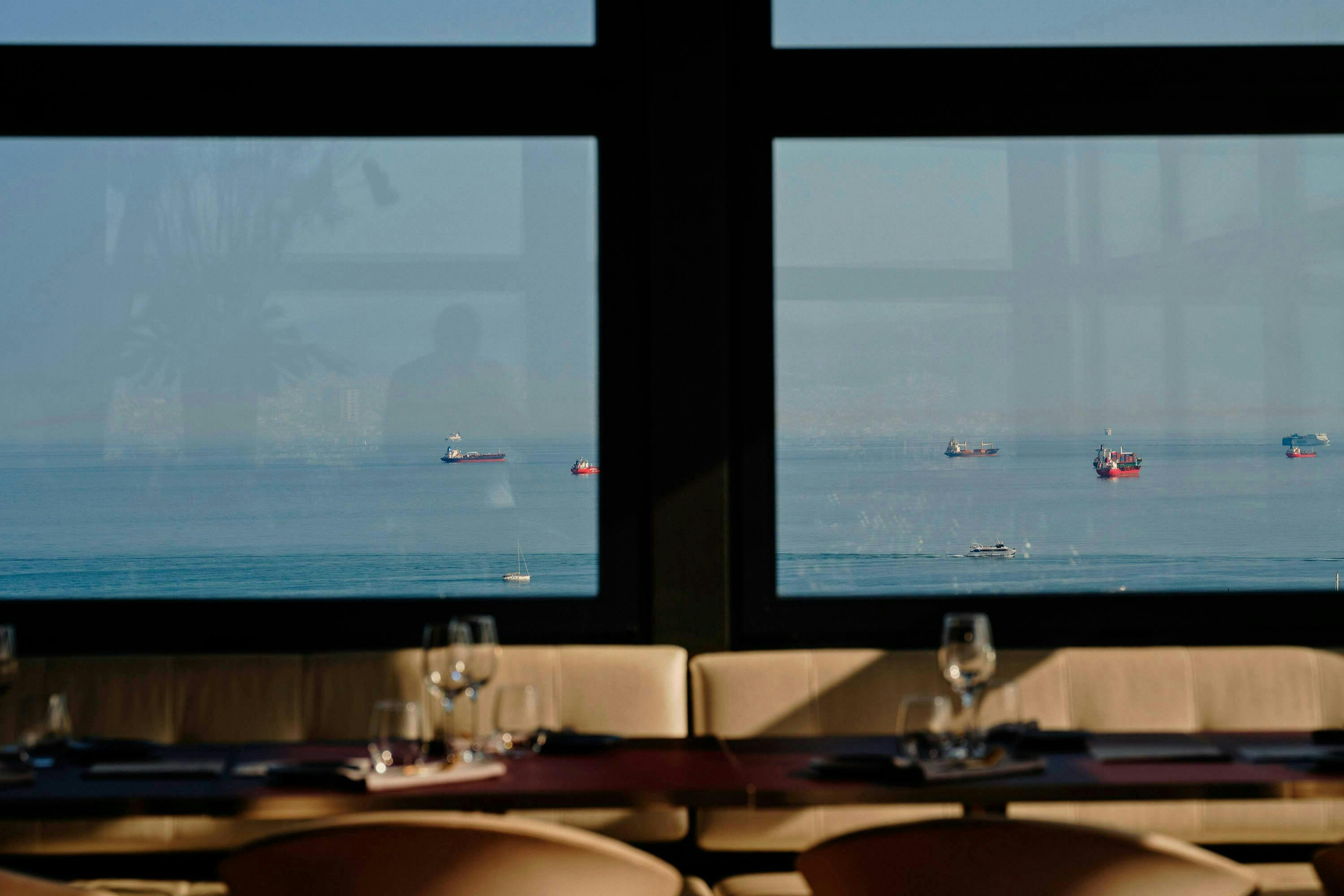 Utsikt över lastfartyg på havet genom ett restaurangfönster med dukade matbord och stolar i förgrunden.