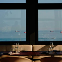 Utsikt över lastfartyg på havet genom ett restaurangfönster med dukade matbord och stolar i förgrunden.