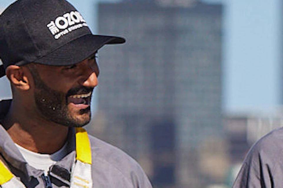Three people in gray jumpsuits and harnesses walk along a high platform with a city skyline in the background. One person points ahead.