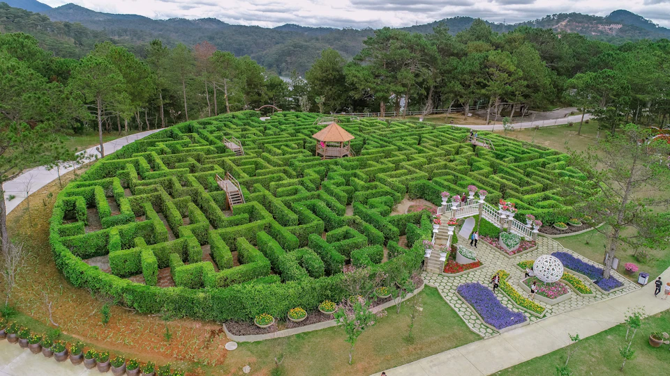 A hedge maze with a central gazebo, surrounded by trees and hills, with paths and decorative elements visible.
