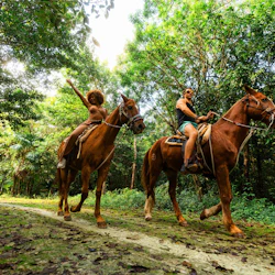 Two people riding horses on a forest trail surrounded by lush greenery, with one person raising their arm joyfully.