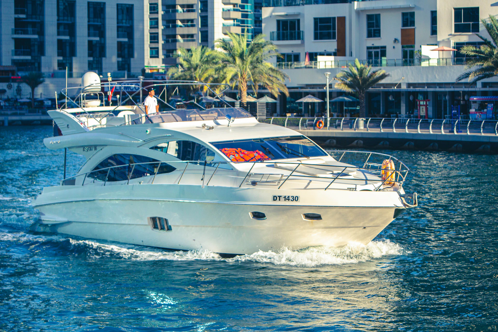 A white yacht cruises through a marina with modern buildings and palm trees in the background.
