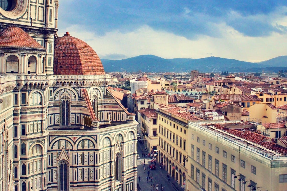 Vue d'un paysage urbain historique comprenant une grande cathédrale avec un dôme en tuiles rouges et une façade complexe, avec des collines lointaines sous un ciel nuageux.