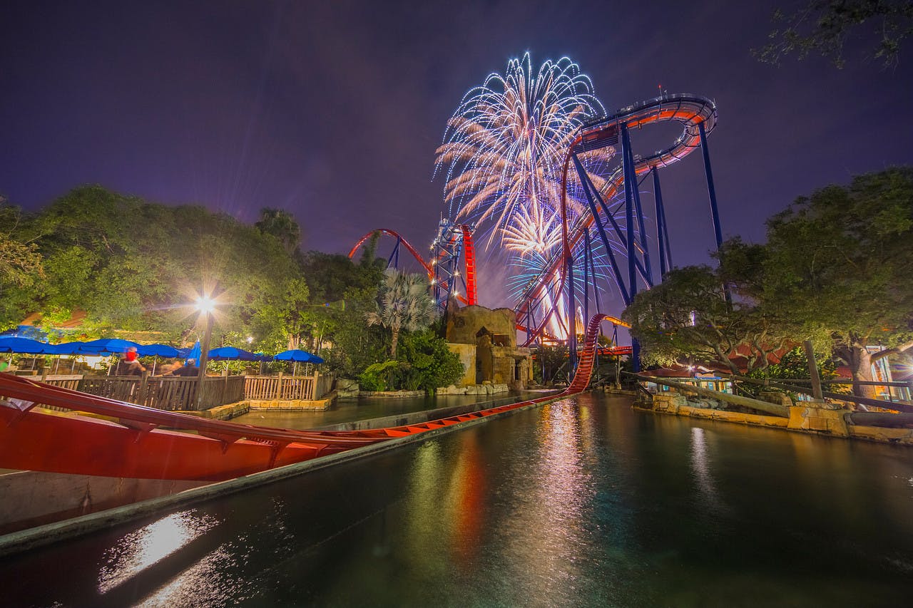 夜には明るく照らされたジェットコースターの上で花火が爆発し、木々や構造物に囲まれた水に反射する軌跡があります。