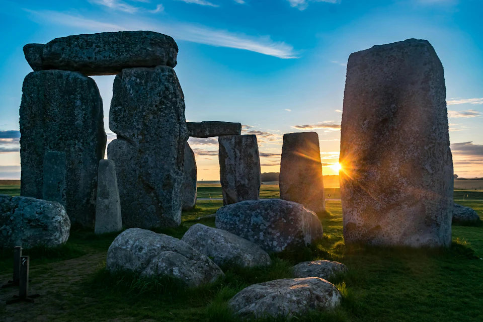 Large stone monoliths with rough surfaces standing upright in a grassy area, illuminated by a bright sunset.