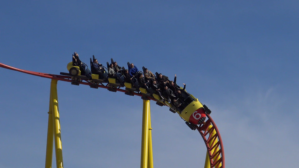 People riding a yellow and red rollercoaster, descending a steep curve against a clear blue sky.