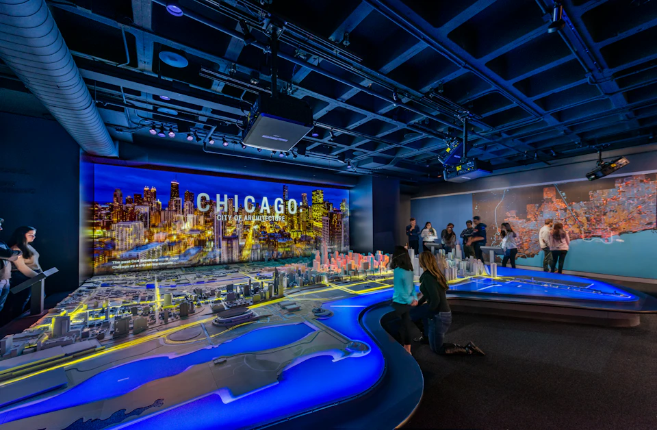 People looking at a detailed illuminated model of Chicago's cityscape in a dark exhibit room with "CHICAGO" lit up in the background.