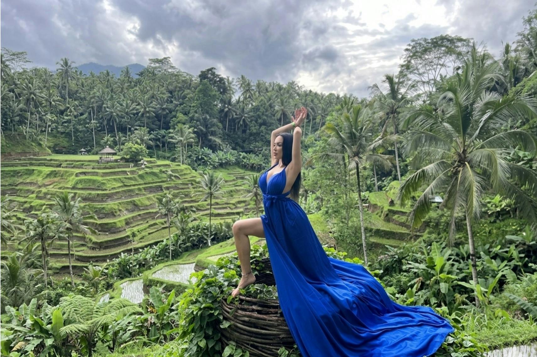 A woman in a flowing blue dress poses gracefully on a scenic terrace surrounded by lush greenery and rice paddies.