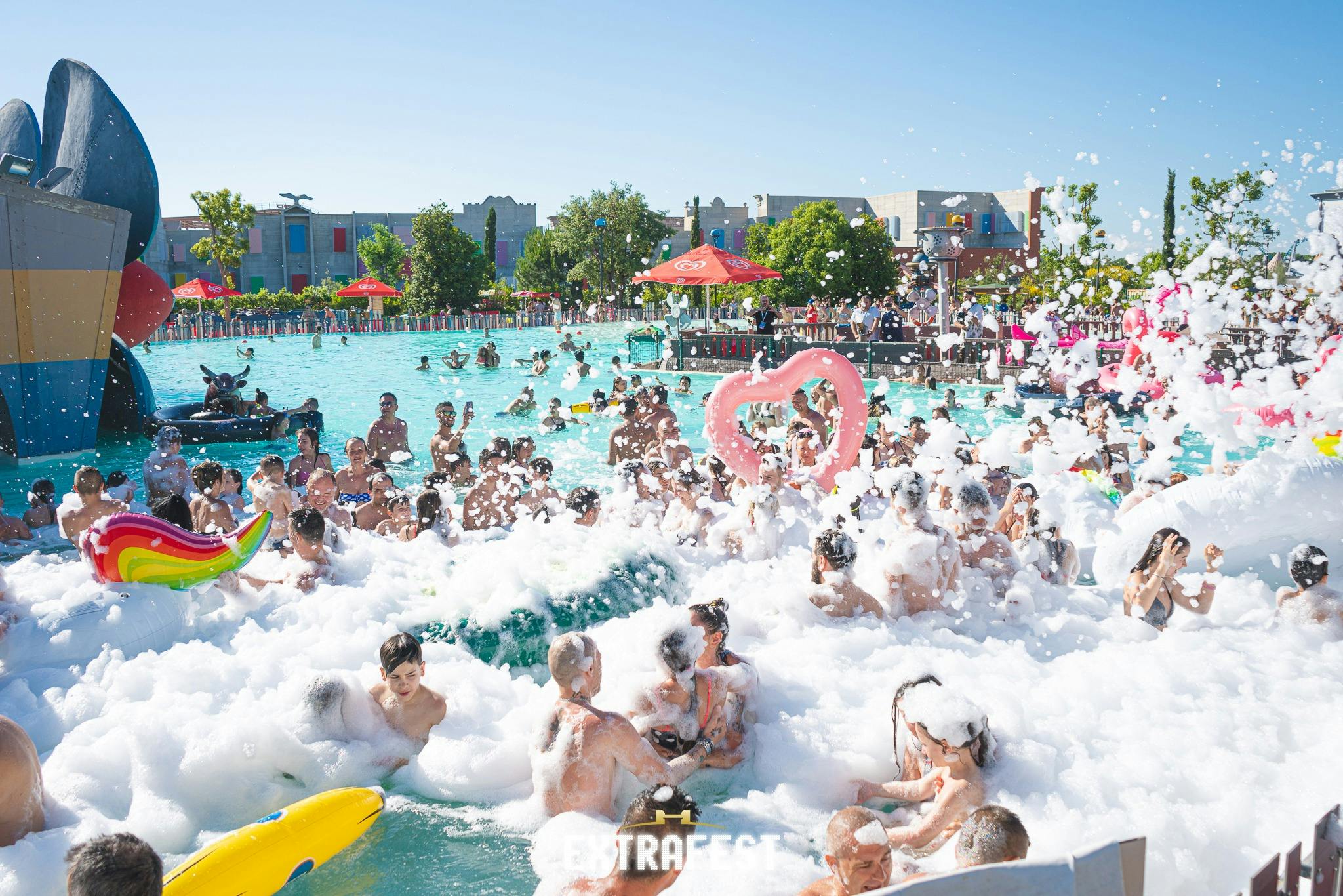 Crowded pool party with people in swimwear; foam bubbles, inflatables, and sun umbrellas are visible under a clear blue sky.