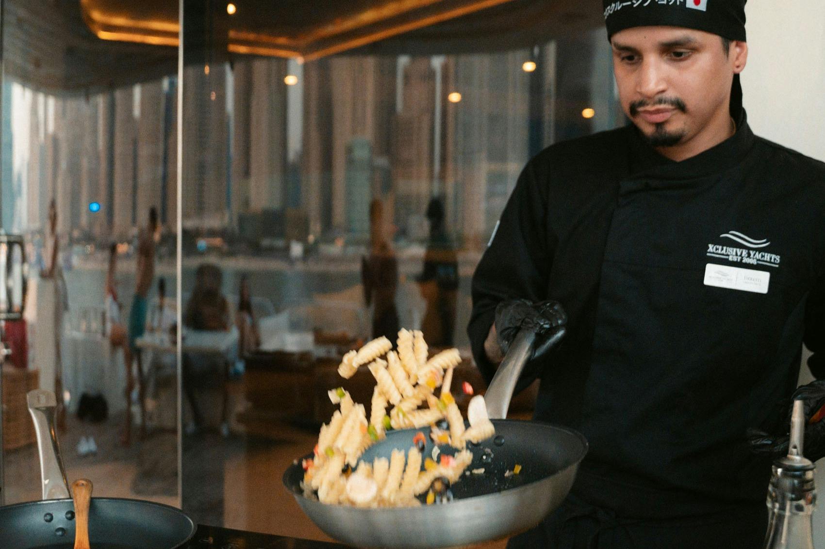 A chef in black attire cooking with a frying pan, tossing crinkle-cut fries and vegetables in a modern indoor setting.